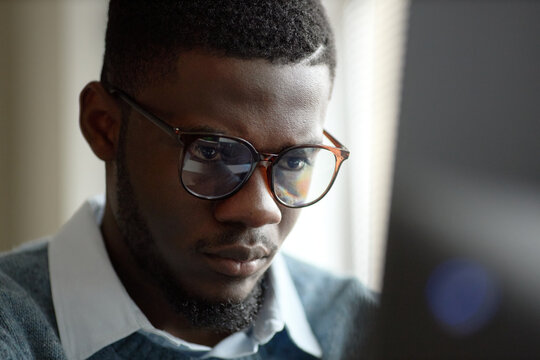 Close up of young adult African American man wearing glasses, focusing on computer screen while working. Ideal for office work, digital productivity, tech career content