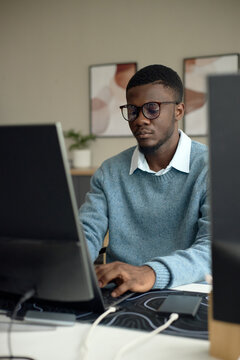 Vertical shot of young adult African American man working at desktop computer, typing and focusing on screen in office setting. Useful for business productivity, remote work, tech career