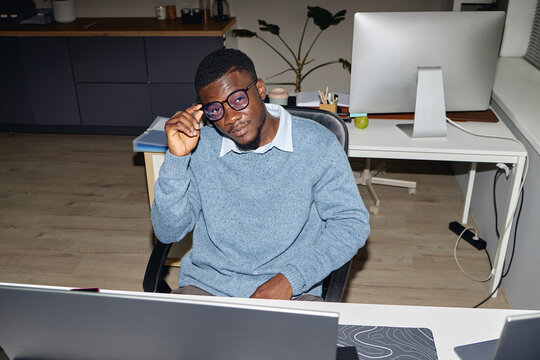 Portrait of young adult Black man adjusting eyeglasses while sitting at office desk, representing focused workplace productivity. Ideal for business, career, corporate content