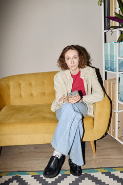 Portrait of young adult Caucasian woman sitting on sofa, holding smartphone, looking at camera, representing mobile communication. Vertical shot for app ads