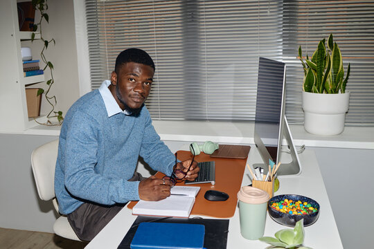 Portrait of young Black man working at desk, writing notes beside desktop computer in office. Supporting office work, productivity or freelance business use