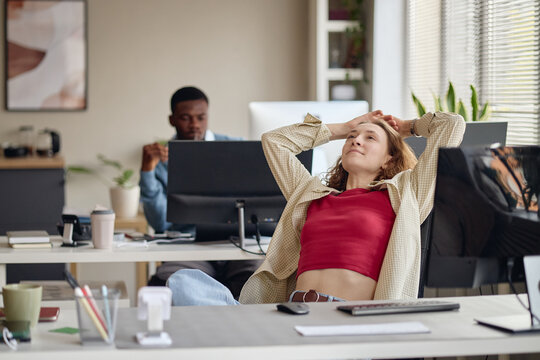 Young adult Caucasian woman leaning back at office desk, stretching during work break while male coworker working in background. Useful for burnout, stress relief, workplace wellness marketing