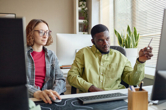 Young adult Caucasian woman and Black man collaborating at desktop computer, reviewing screen content and discussing workflow. Ideal for teamwork, office training, productivity marketing