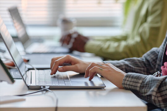 Close up of young adult coworkers typing on laptops at shared desk, collaborating on digital tasks in office. Ideal for teamwork, productivity, workplace technology marketing