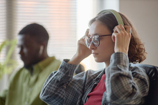 Young adult Caucasian woman adjusting headphones while working beside Black man in office. Depicting focused teamwork and audio communication for workplace training or tech support