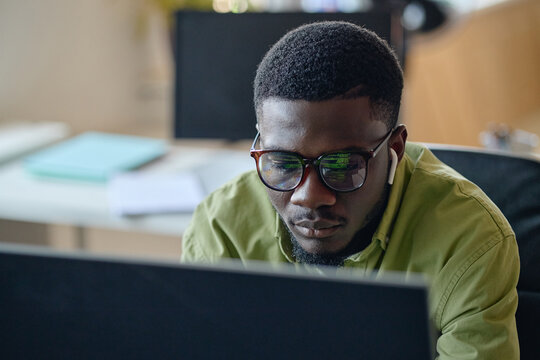 Close up of young adult African American man working on computer, wearing earbuds and focusing on screen. Ideal for remote work, coding, productivity, digital business content