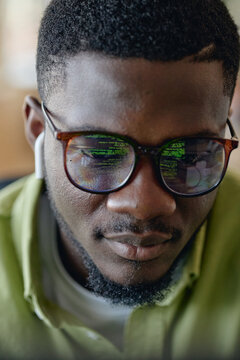 Vertical shot of Black young adult man coding on computer, reflecting data on glasses and wearing earbuds. Representing software development, cybersecurity, tech recruitment marketing