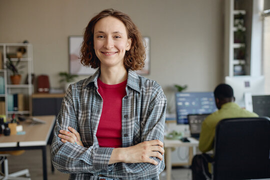 Portrait of young adult Caucasian woman standing with crossed arms in office, smiling at camera while coworker working in background. Ideal for workplace culture, team leadership, recruitment