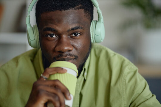 Portrait of young adult Black man wearing headphones and drinking coffee while looking at camera, suggesting podcast listening or remote work break. Close up for audio apps or lifestyle marke