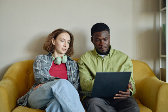 Young adult Caucasian woman and Black man reviewing laptop together on sofa, discussing online project and teamwork. Useful for remote work, startup collaboration, digital learning