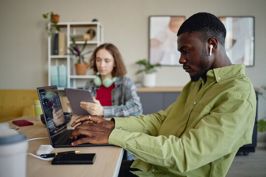 Young Black man working on laptop while Caucasian woman using tablet in shared office, showing teamwork and digital productivity. Ideal for remote work, startup, collaboration marketing
