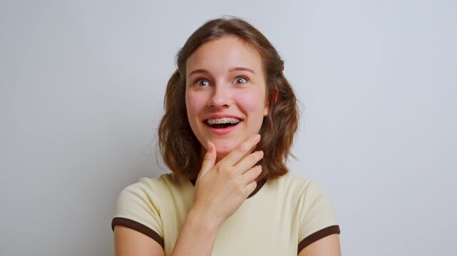 young woman looking surprised and shocked, covering her mouth in disbelief and astonishment with her hand against a simple studio background