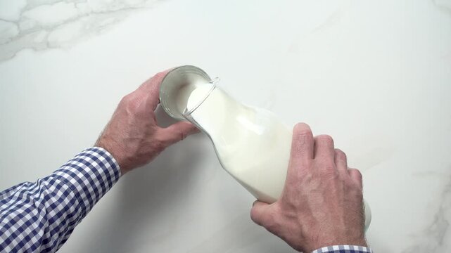 Man's hand pours fresh milk from a glass bottle into a clear glass on a white marble table. Symbolizes dairy nutrition, healthy breakfast, and organic farming. Ideal for food and wellness content