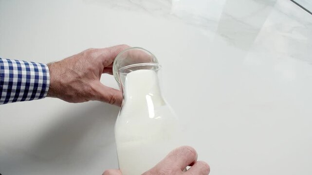 Man's hand pours fresh milk from a glass bottle into a clear glass on a white marble table. Symbolizes dairy nutrition, healthy breakfast, and organic farming. Ideal for food and wellness content