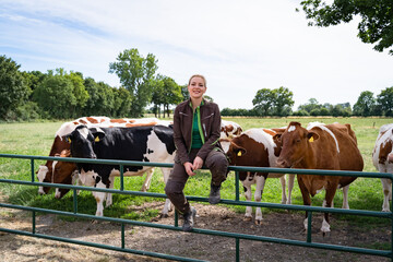 A smiling young female farmer in workwear sits on a green metal pasture gate, with a group of black-and-white and red-and-white dairy cows standing on the meadow behind her. © Countrypixel