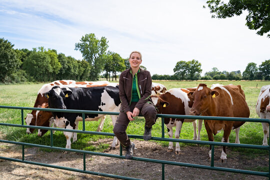 A smiling young female farmer in workwear sits on a green metal pasture gate, with a group of black-and-white and red-and-white dairy cows standing on the meadow behind her.