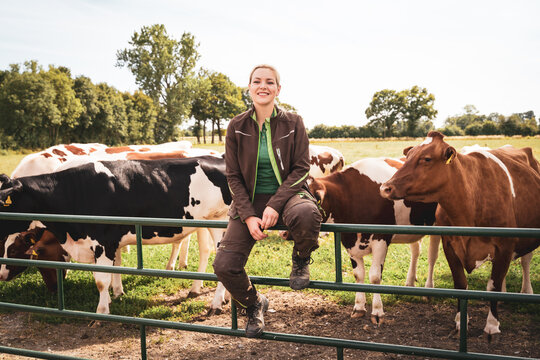 Eine l&auml;chelnde junge Landwirtin in Arbeitskleidung sitzt auf einem gr&uuml;nen Metall-Weidetor, w&auml;hrend im Hintergrund eine Gruppe von schwarz-bunten und rot-bunten Milchk&uuml;hen auf der Koppel steht.