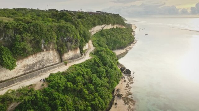 Lush green cliffs cascading down to pristine sandy beach and clear turquoise ocean water, featuring boats anchored offshore and a winding road along the dramatic coastline of Bali. Aerial drone flight