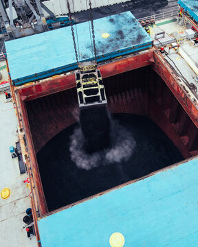 Aerial view of a crane grab unloading black coal into the cargo hold of a ship at an industrial port in Riga, Latvia.