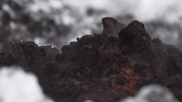 Dark charred rock formations with rough porous texture and soft gray haze in close macro view, creating gritty abstract industrial atmosphere with muted black, rust, and charcoal tones