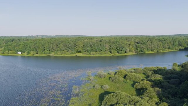 Drone footage capturing a wide aerial view of Lac du Bourdon in France, showing the calm lake bordered by dense green forest and lush vegetation. The scene is tranquil with clear blue skies and no