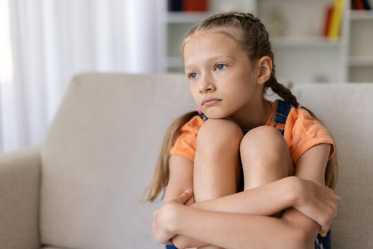 Pensive teen girl sitting alone on sofa at home hugging her knees, showing sadness, loneliness, boredom or emotional stress in a quiet lifestyle portrait