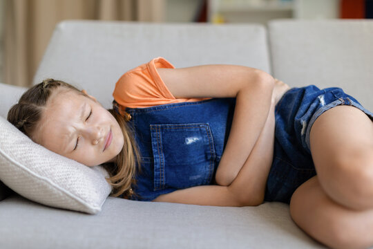Teen girl lying on sofa at home holding her stomach with eyes closed, showing abdominal pain, period cramps or illness in child healthcare lifestyle scene