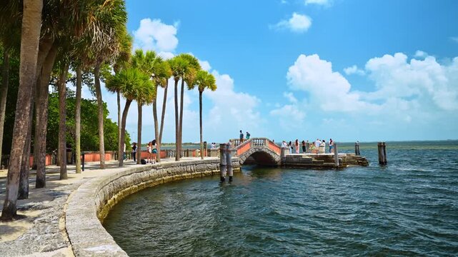 Miami, USA, 16 April 2026: Visitors on stone bridge at Vizcaya bayfront, Miami, Florida. Multiple people on ornamental pier with palm trees, seawall and choppy water under blue sky.