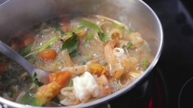 Spoon stirring Thai Tom Yum Goong hot & sour soup, simmering shrimp prawns & aromatic lemongrass, kaffir lime leaves herbs vegetables in boiling cooking pot. Asian home cooked meal
