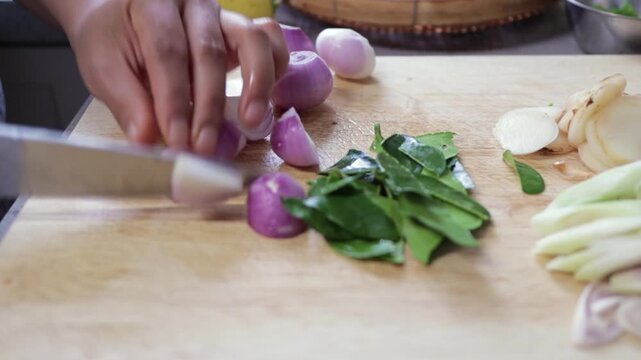 Hands slice chop organic fresh raw shallots on wood cutting board. Preparation of aromatic Thai Asian ginger root, kaffir lime leaf herb, & lemongrass, authentic Thai home cooking