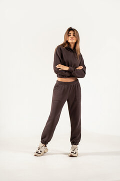 Full-length portrait of a confident young woman wearing a trendy brown oversized tracksuit and chunky sneakers, standing with crossed arms in a minimalist studio against a white background. 
