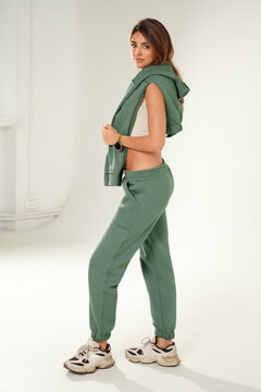 Full-length portrait of a confident young woman wearing a trendy tracksuit and chunky sneakers, standing  in a minimalist studio against a white background.