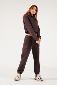 Full-length portrait of a confident young woman wearing a trendy brown oversized tracksuit and chunky sneakers, standing with crossed arms in a minimalist studio against a white background