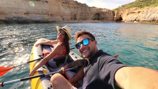 Happy young couple taking a selfie video with a wide-angle camera while enjoying a kayak excursion on a sunny day near a beautiful coastline with impressive cliffs and sea caves