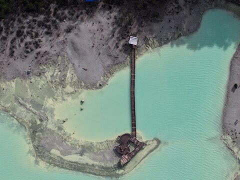 Aerial view of the turquoise waters of Kawah Putih with a wooden walkway extending into the lake, surrounded by volcanic terrain in Kecamatan Ciwidey, Jawa Barat, Indonesia.