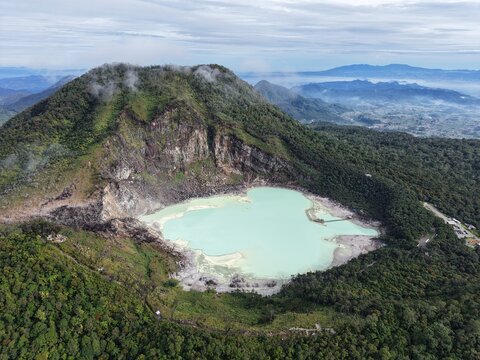 Aerial view of the turquoise volcanic lake of Kawah Putih surrounded by lush green forest and rugged crater walls in Kecamatan Ciwidey, Jawa Barat, Indonesia.