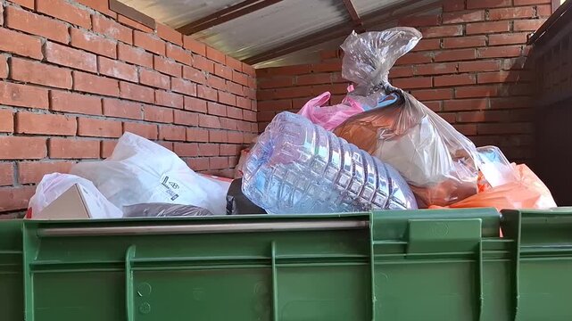 Person throwing garbage bag into crowded green dumpster outdoors. Municipal garbage disposal, recycling issues, environmental pollution. Large dumpster with plastic bottles. Ecology, waste management