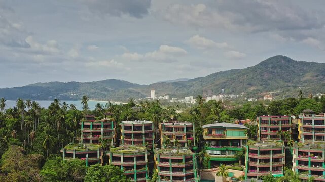 Tropical luxury resort community buildings nestled on green hillside, surrounded by palm trees and mountains overlooking ocean in Phuket, Thailand. Summer vacation accommodations. Aerial drone flight