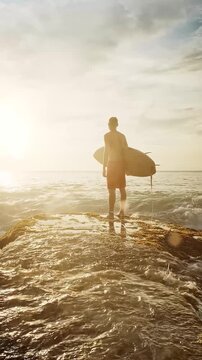 Young surfer stands in the ocean at sunset, holding his surfboard and savoring a tranquil moment before catching the waves. Warm golden hour light reflects off the water, highlighting the serene