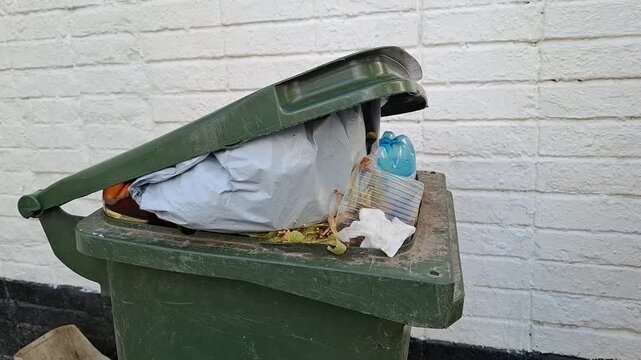 Person throwing crumpled white paper napkin into overflowing green dumpster outdoors. Municipal garbage disposal, recycling issues, environmental pollution. Ecology, social issues, waste management