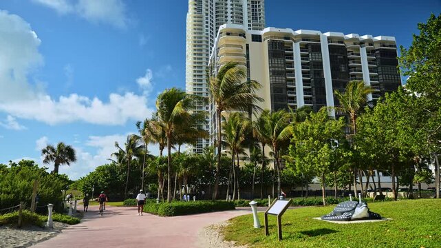 Miami, USA, 16 April 2026: Cyclists on beachside boardwalk near condominiums, Sunny Isles Beach, Florida. Red paved path with palms, sculpture and green lawn on a sunny day.