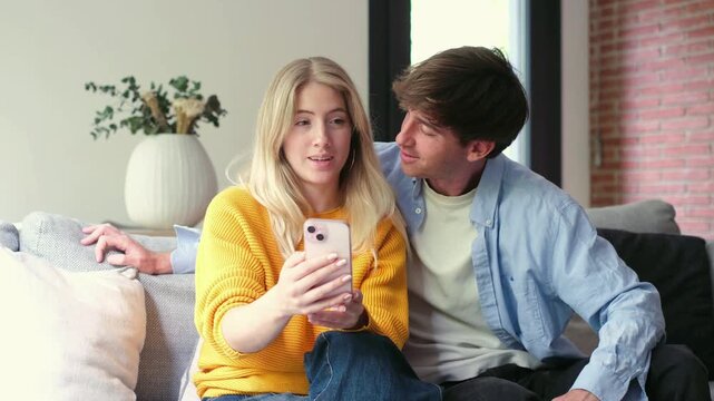 Young caucasian couple sitting on a couch in their living room, happily browsing social media, watching videos, or making a video call on a smartphone together, sharing a moment of connection
