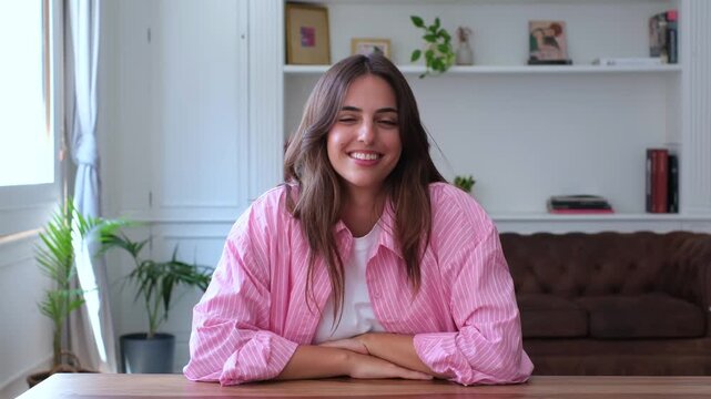 Pretty young caucasian female wearing casual clothes sitting at a wooden desk at home. Confident student smiling and looking at camera during an online lesson or job interview