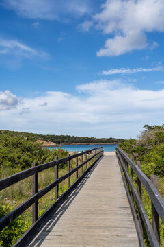 View of Spiaggia del Relitto with a wooden boardwalk leading to the turquoise sea, surrounded by Mediterranean scrub under a blue sky in La Maddalena, Sardinia, Italy.