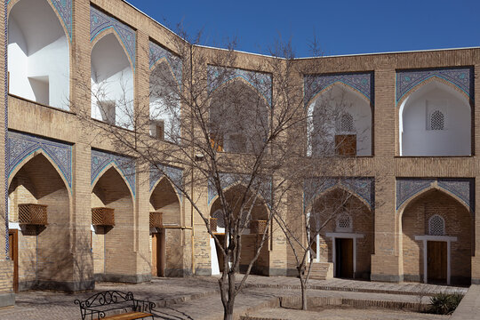 Courtyard of Muhammad Amin Khan Madrasah in Khiva with pointed arches and blue tilework. Historic two-story brick facade with decorative Islamic patterns surrounding a tranquil empty courtyard.