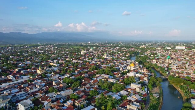 Aerial drone footage of Mataram city, Lombok island, Indonesia, with many local houses, many beautiful mosques, vegetations, and mountains in the background, at sunset