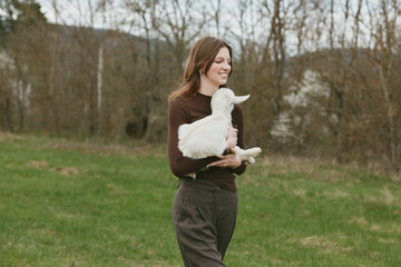 Obraz premium Smiling woman holds a goat kid in a grassy field, enjoying an outdoors moment with a calm rural feel, highlighting animal human bond and nature.