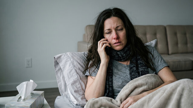Miserable woman with a cold making a phone call while wrapped in a blanket at home.