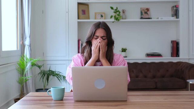 Desperate young woman sitting at a desk in her living room, reacting with shock and sorrow after reading bad news on her laptop, eventually covering her face and starting to cry