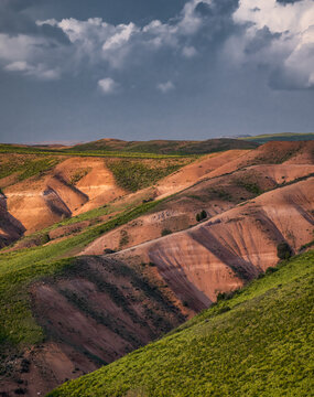 View of the rolling hills of Arashan with red earth slopes, patches of green vegetation, and dramatic storm clouds in the sky, Pop District, Namangan Region, Uzbekistan.